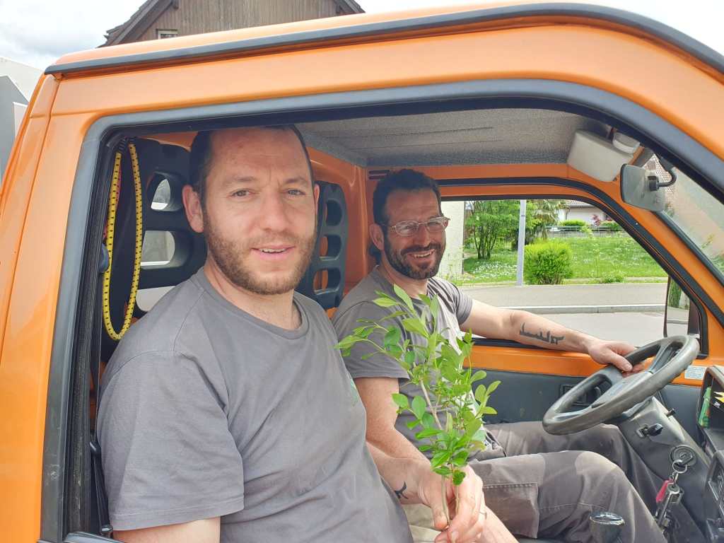 Zwei Männer sitzen in einem orangefarbenen Lieferwagen. Der Mann auf der linken Seite hält einen kleinen Zweig, während der andere lächelt und die Steuerung in der Hand hat.